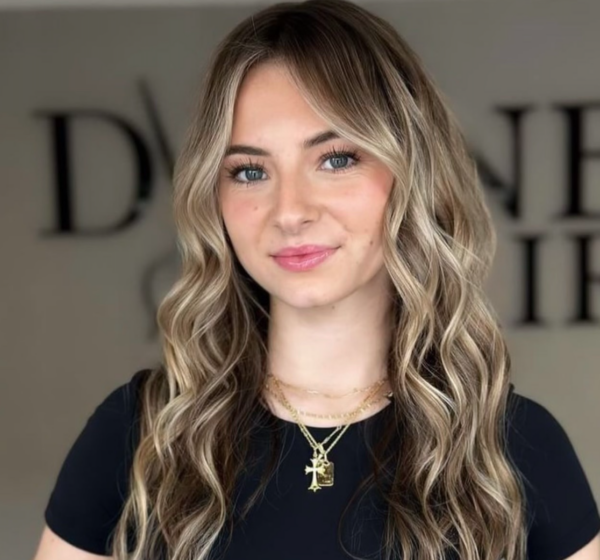 Define Hair & Skin in Ellicott City, MD A woman with lived-in color, long wavy blonde hair, and blue eyes, wearing a black top and layered necklaces, stands in front of a blurred background with indistinct text.