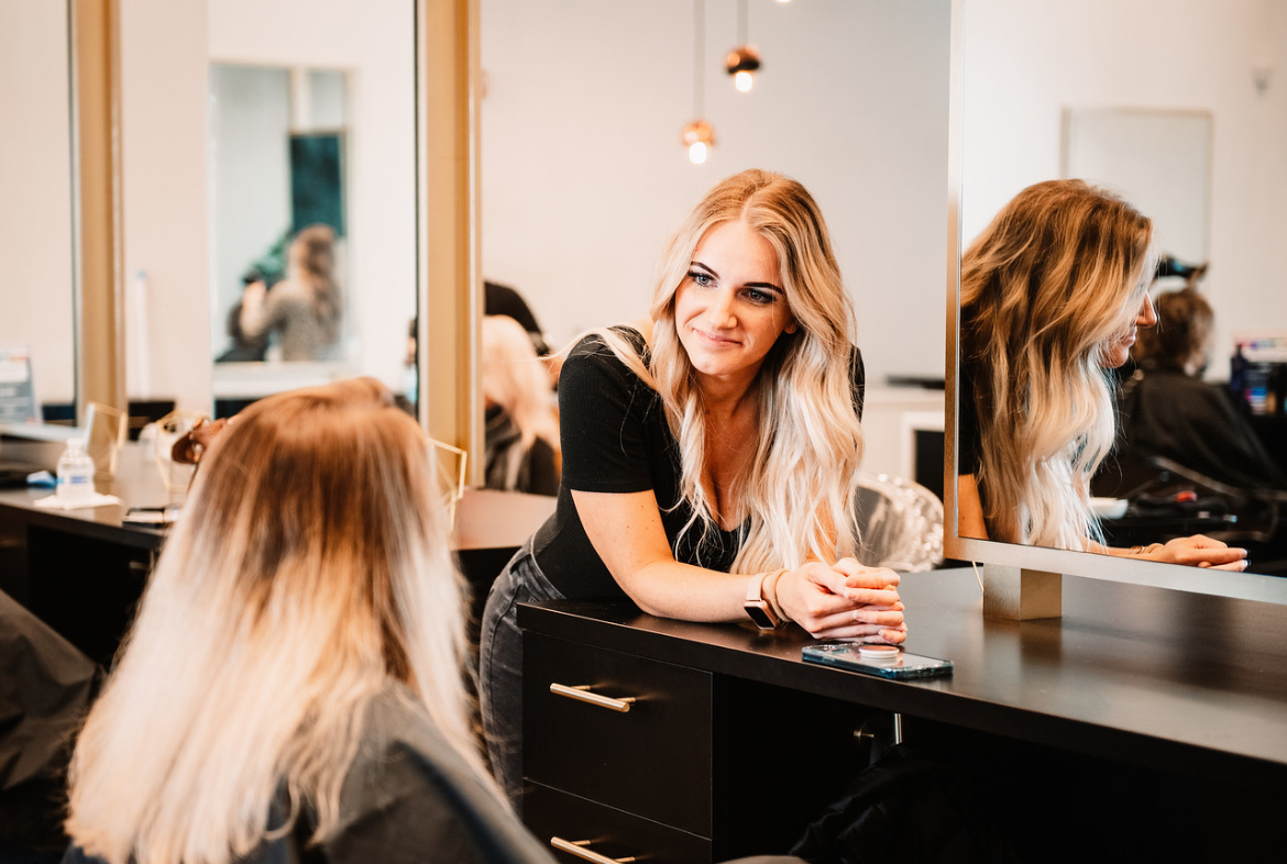 Define Hair & Skin in Ellicott City, MD A woman with long blonde hair is looking at another woman in a hair salon. During your first visit, both are near a mirror, and the blonde woman is leaning on a counter with a phone beside her.