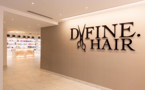 Define Hair & Skin in Ellicott City, MD Entrance of a hair salon with a wall sign "DVFINE HAIR" featuring scissors, and a well-lit reception desk displaying hair products in the background. On your first visit, get defined with expert hairstyles and premium products.