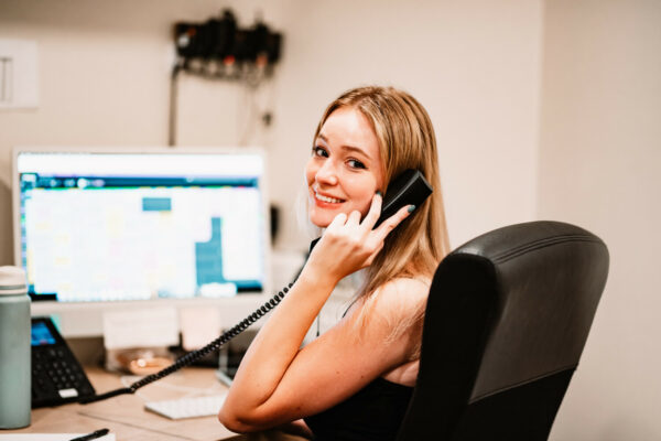 Define Hair & Skin in Ellicott City, MD A woman sits at a desk, smiling while talking on a telephone during your first visit. A computer screen with colorful charts and a water bottle are visible in front of her.