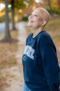 Define Hair & Skin in Ellicott City, MD A person with a shaved head, smiling and looking up, stands on a tree-lined sidewalk wearing a dark sweatshirt, embodying the essence of glowing up.